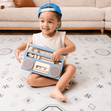 Baby toddler sitting and holding a toy radio while smiling on the toddlekind baby toddler foam modern designer puzzle play mat haven amira seagrass.