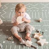 Baby sitting and chewing toy while surrounded by wooden toys on toddlekind baby toddler foam modern designer puzzle play mat haven berber moss.