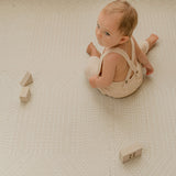 Baby sitting and looking back while playing with wooden blocks on toddlekind baby toddler foam modern designer puzzle play mat haven deco ecru