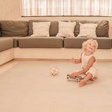 Toddler sitting and smiling with toys on the toddlekind baby toddler foam modern designer puzzle play mat haven deco latte in a bright living room.