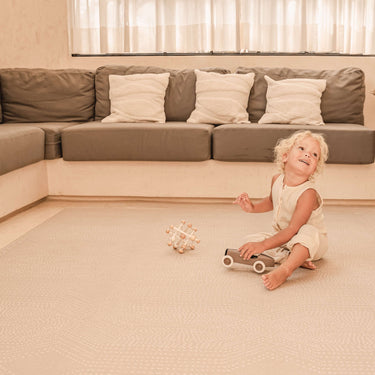Toddler sitting and smiling with toys on the toddlekind baby toddler foam modern designer puzzle play mat haven deco latte in a bright living room.