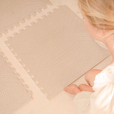 Person assembling interlocking tiles of the toddlekind baby toddler foam modern designer puzzle play mat haven deco latte on a light floor.