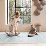 Toddler standing on a rocking toy while baby sits nearby on the toddlekind baby toddler foam modern designer puzzle play mat haven linear mineral.