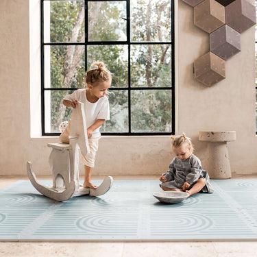 Toddler standing on a rocking toy while baby sits nearby on the toddlekind baby toddler foam modern designer puzzle play mat haven linear mineral.