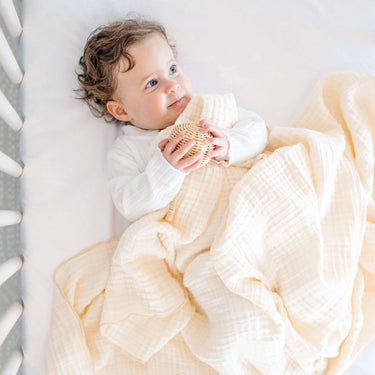 Baby lying comfortably with toddlekind 3 layer cotton muslin baby blanket kind cloth blush draped gently over body.