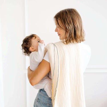 Parent holding baby wrapped in toddlekind 3 layer cotton muslin baby blanket kind cloth sage in a bright minimal home.