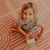 Smiling toddler playing with a wooden toy while sitting on the toddlekind baby toddler foam modern designer puzzle play mat haven linear camel.