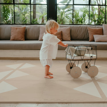 Baby standing and pushing toy stroller across toddlekind baby toddler foam modern designer puzzle play mat haven kyte mocha in a bright living room.