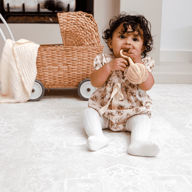 Baby toddler sitting on the mat holding a woven toy with a wicker cart nearby, showcasing the toddlekind baby toddler foam modern designer puzzle play mat haven parisa millet.