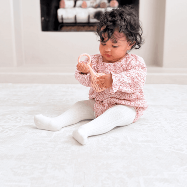 Toddler seated and examining a toy while sitting on the toddlekind baby toddler foam modern designer puzzle play mat haven parisa millet in a cozy living space.