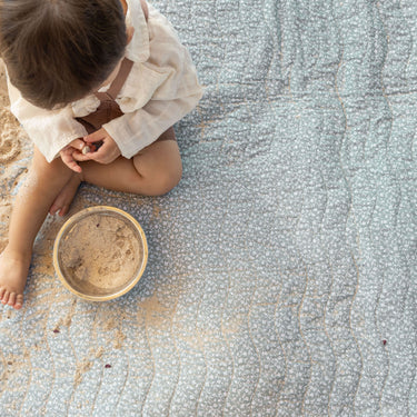 Overhead view of toddler playing with sand and a bowl on a sage green padded mat, illustrating the toddlekind portable water resistant wander classic play mat botanical sage for mess-friendly play.