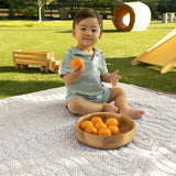 Toddler sitting outdoors on a floral mat holding an orange with a wooden bowl of fruit nearby, highlighting the toddlekind portable water resistant wander classic play mat botanical earthen in a sunny backyard setting.