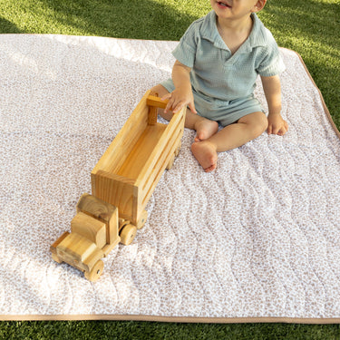 Toddler playing with a wooden toy truck on a padded floral mat in the grass, demonstrating the toddlekind portable water resistant wander classic play mat botanical earthen for comfortable outdoor play.