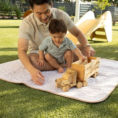 Parent and toddler sitting together on a floral mat playing with a wooden toy truck, highlighting bonding time on the toddlekind portable water resistant wander classic play mat botanical earthen.