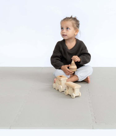 Toddler sitting and playing with wooden train and spinning top on toddlekind baby toddler foam modern puzzle play mat classic stone.