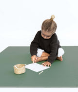 Toddler drawing with crayon while sitting on toddlekind baby toddler foam modern puzzle play mat classic moss with woven basket nearby.