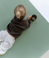 Overhead view of toddler lying on toddlekind baby toddler foam modern puzzle play mat classic moss reaching for snack cup.