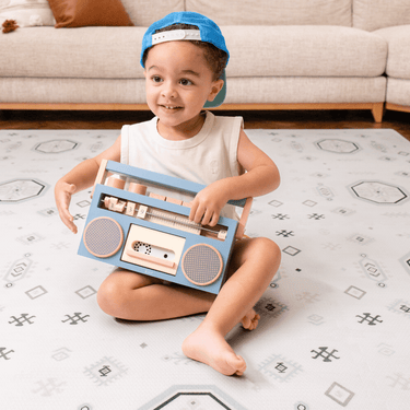 Baby toddler sitting and holding a toy radio while smiling on the toddlekind baby toddler foam modern designer puzzle play mat haven amira seagrass.
