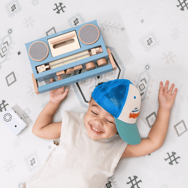 Overhead view of baby toddler lying down with a toy radio, enjoying the toddlekind baby toddler foam modern designer puzzle play mat haven amira seagrass
