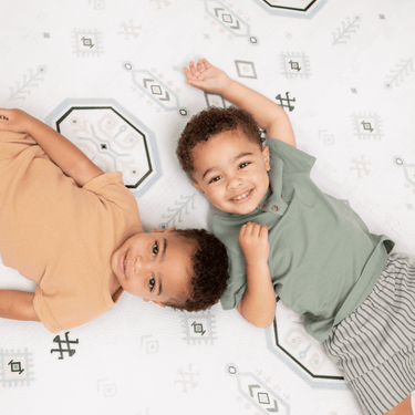 Two baby toddlers lying side by side and smiling on the soft surface of the toddlekind baby toddler foam modern designer puzzle play mat haven amira seagrass.
