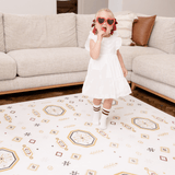 Baby toddler standing in a living room wearing heart sunglasses and enjoying a treat on the toddlekind baby toddler foam modern designer puzzle play mat haven amira golden clay.
