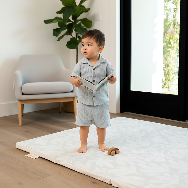 Toddler standing and playing in living room on toddlekind baby toddler foldable foam cushioned tumble play mat willow frost.

