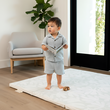 Toddler standing and playing in living room on toddlekind baby toddler foldable foam cushioned tumble play mat willow frost.
