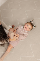 A smiling toddler lies on tulum desert caliza interlocking foam play mat with a subtle geometric pattern while playing with a caregiver. The cushioned play mat provides a comfortable, supportive surface for active play and bonding moments.