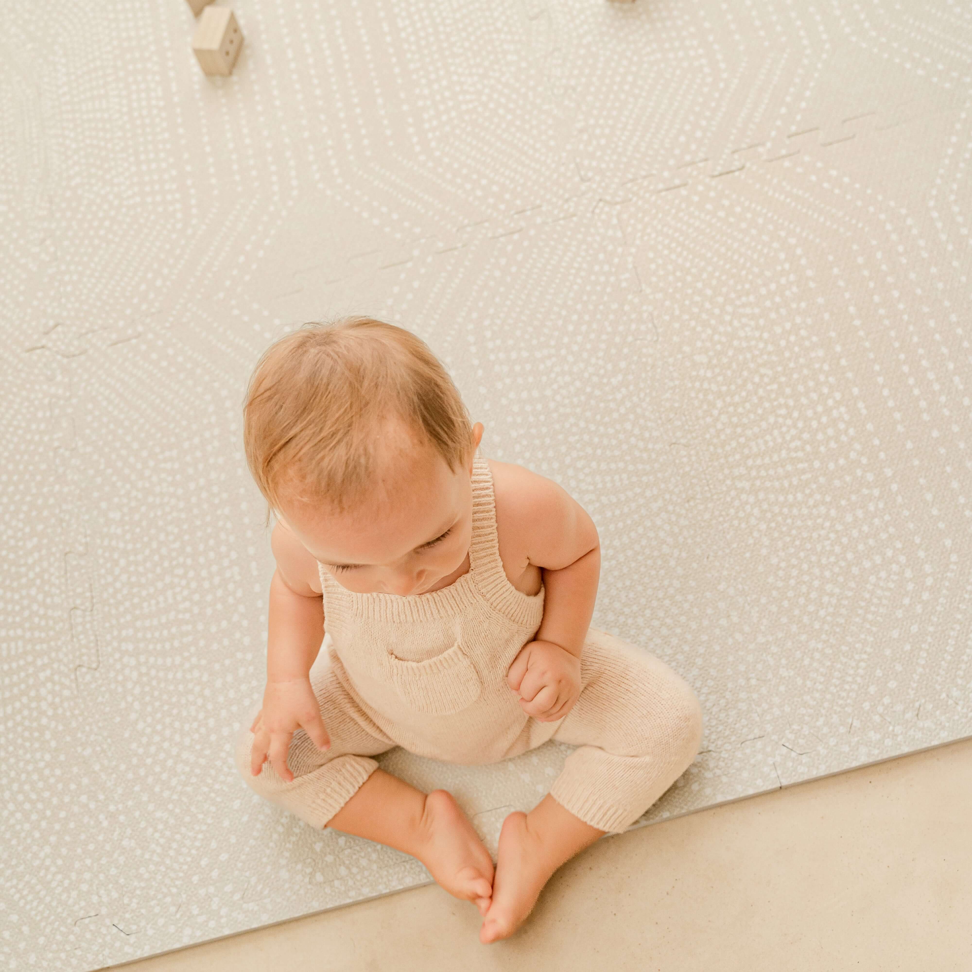 Baby sitting on the toddlekind baby toddler foam modern designer puzzle play mat haven deco ecru wearing neutral knit outfit.
