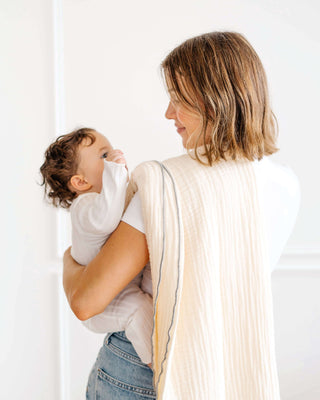 Parent holding baby wrapped in toddlekind 3 layer cotton muslin baby blanket kind cloth sage in a bright minimal home.
