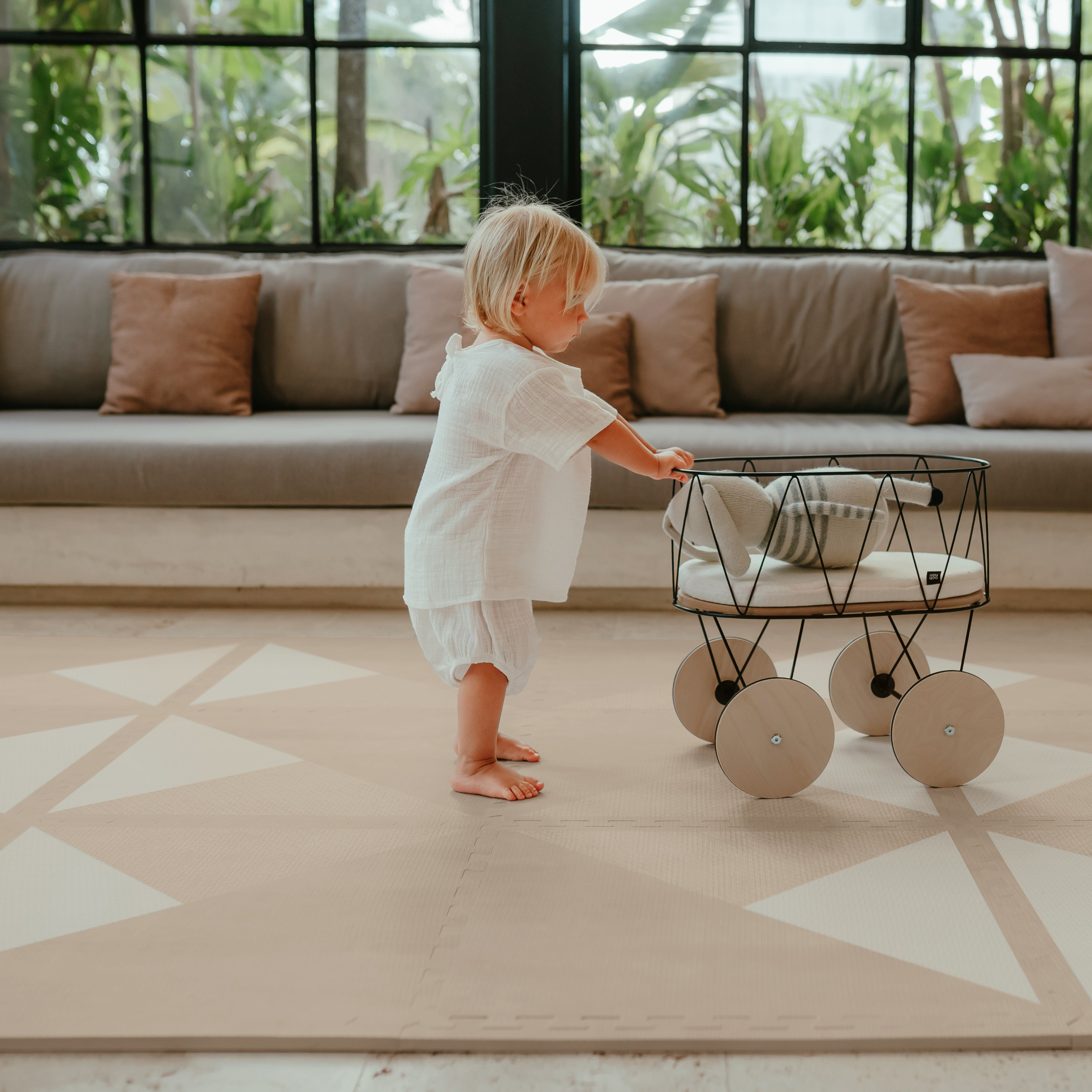 Baby standing and pushing toy stroller across toddlekind baby toddler foam modern designer puzzle play mat haven kyte mocha in a bright living room.
