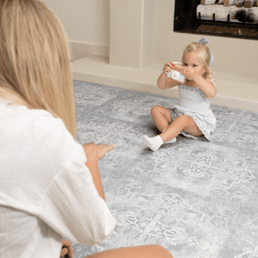 Toddler sitting on the mat holding a toy camera while parent interacts nearby, illustrating everyday play on the toddlekind baby toddler foam modern designer puzzle play mat haven parisa pearl blue.

