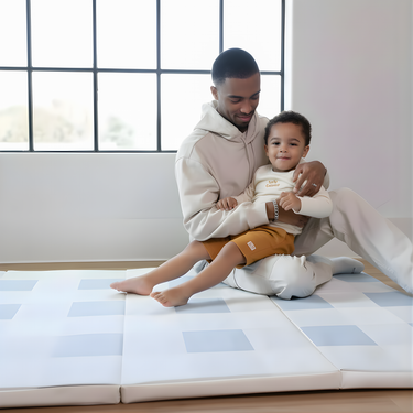 Father and toddler sitting together on toddlekind baby toddler foldable foam cushioned tumble play mat arden tide in bright room.
