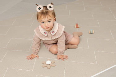 A baby sits on a soft beige interlocking foam play mat with a subtle geometric pattern, playing with wooden toy cars and a flowertoy. The cushioned mat provides a comfortable, safe surface for crawling and early play.