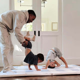 Parent helping toddler play and stretch on toddlekind baby toddler foldable foam cushioned tumble play mat arden tide.
