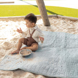 Baby sitting on a soft sage green mat in a sandbox, playing with sand and a small bowl, showcasing the toddlekind portable water resistant wander classic play mat botanical sage in an outdoor setting.
