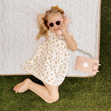 Young child lying on a floral mat wearing sunglasses and holding a toy phone, illustrating playful moments on the toddlekind portable water resistant wander classic play mat botanical earthen outdoors.
