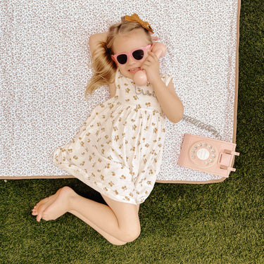 Young child lying on a floral mat wearing sunglasses and holding a toy phone, illustrating playful moments on the toddlekind portable water resistant wander classic play mat botanical earthen outdoors.
