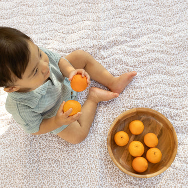 Overhead view of a baby playing with oranges on a soft floral mat with a wooden bowl, showcasing the toddlekind portable water resistant wander classic play mat botanical earthen for sensory play.
