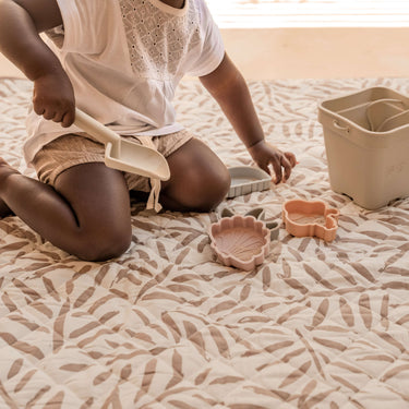 Close-up of toddler scooping sand with toys on cushioned mat – toddlekind portable water resistant wander classic play mat ocean tan for mess-free play
