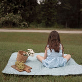 Child sitting on a sage green floral mat during a picnic with grapes, flowers, and a basket, highlighting the toddlekind portable water resistant wander classic play mat botanical sage for outdoor relaxation.
