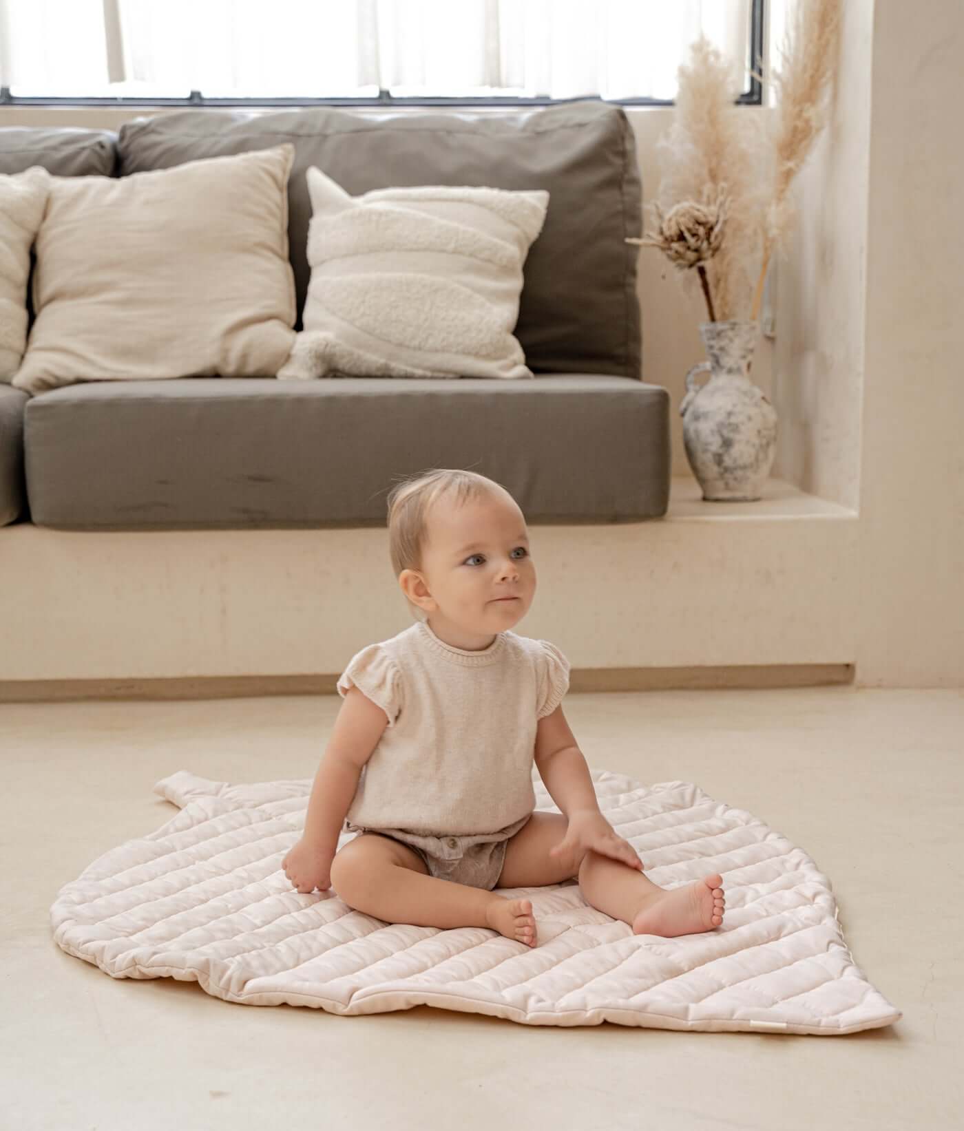 Baby sitting on organic cotton padded baby toddler toddlekind play mat tummy time quilted leaf mat blush in a soft neutral living room.
