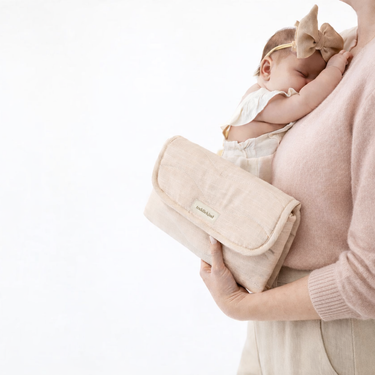 Parent holding folded toddlekind baby portable changing clutch mini maven blush with baby resting against shoulder.
